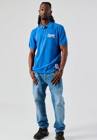 Blauwe poloshirt met wit logo op de borst, in combinatie met lichtblauwe jeans en zwart camouflage sneakers, staand tegen een neutrale achtergrond.