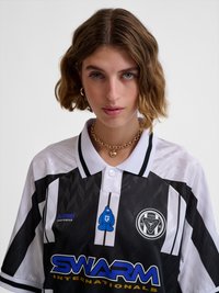 Junge Person mit kurzen, gewellten braunen Haaren trägt ein schwarz-weißes Sporttrikot mit "SWARM"-Logo, eine goldene Halskette und goldene Creolen.