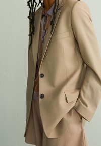 Blazer marron clair en tissu lisse, avec une seule poche, deux boutons foncés, et une chemise à motifs contrastés en dessous.