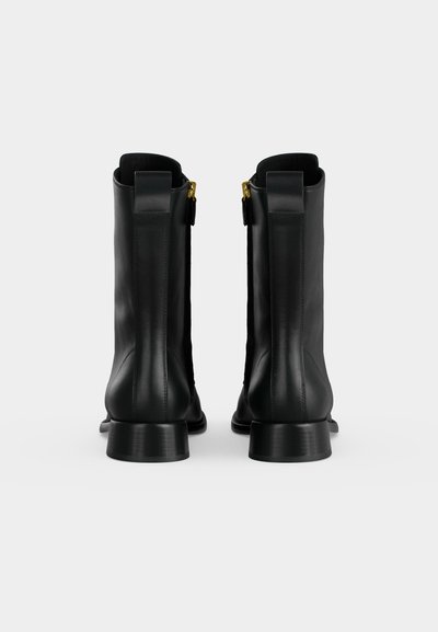 Paire de bottines en cuir noir vues de dos avec tirettes de fermeture éclair dorées et petits talons blocs sur fond uni clair.