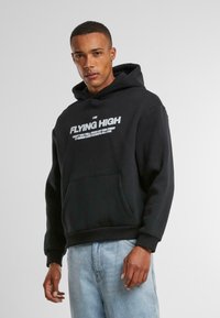 Felpa nera con cappuccio e tasca frontale, presenta una scritta bianca "I am FLYING HIGH" e uno slogan più piccolo. Realizzata in un tessuto morbido, vestibilità rilassata.