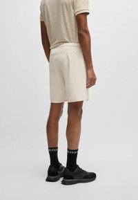 Pantalones cortos color beige claro con un acabado suave, que cuentan con una cinturilla elástica. Llevados con zapatillas negras y calcetines que muestran un logo.