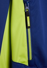 Blå och limegrön jacktyg med en dragkedjeficka med kontrasterande dragkedja, slät textur och färgblockeringsdesign.