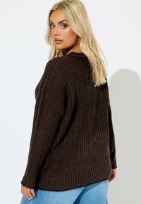 Brauner, strukturierter Strickpullover mit losem Schnitt und langen Ärmeln, mit einem runden Ausschnitt und gerippten Bündchen, getragen über hellblauen Jeans.