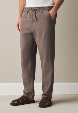 Homme portant un pantalon décontracté marron clair à cordon et des sandales marron foncé à bout ouvert, debout contre un mur neutre.