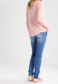Blouse rose clair à manches longues avec un design fluide, associée à un jean ajusté bleu clair. La tenue est complétée par des chaussures plates beiges.