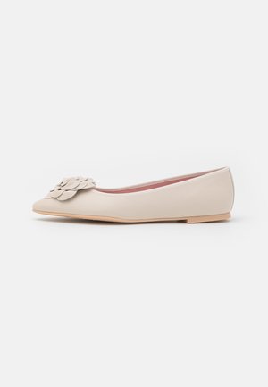 Beige damesko med flad hæl og spids tå, blød sål og lagdelt blomstret dekoration foran, vist i sideprofil mod hvid baggrund.