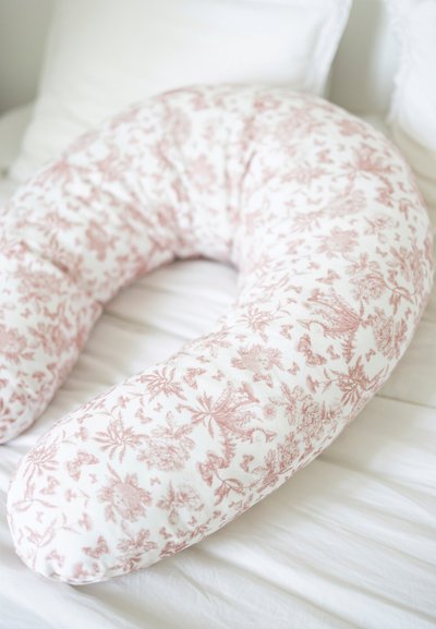 Καμπυλωτό μαξιλάρι σώματος καλυμμένο με λευκό ύφασμα που διαθέτει ροζ λουλουδάτο μοτίβο. Μαλακή υφή με στρογγυλεμένες άκρες. Τοποθετημένο σε λευκό κρεβάτι.