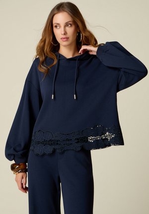Vrouw draagt een marineblauwe hoodie met trekkoorden en kanten zoom, bijpassende broek, grote creolen en bruine armbanden, poserend tegen een effen achtergrond.