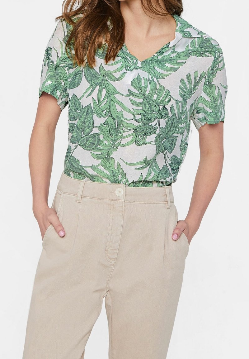 Blusa a maniche corte con motivo di foglie tropicali verdi su sfondo chiaro, con scollo a V e vestibilità rilassata. Abbinata a pantaloni beige.