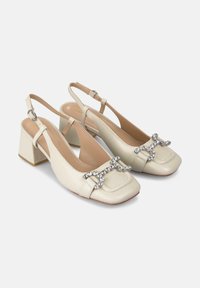Talons slingback en cuir verni beige avec un bout carré, un talon bloc et un détail de boucle en strass décoratif. Texture lisse et design minimal.