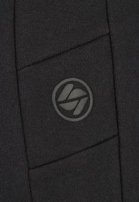 Zwart gebreide stof met een gestructureerd patroon. Voorzien van een grijs cirkelvormig logo met een abstract ontwerp, geplaatst aan de linkerkant.