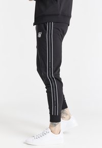 Black athletic joggers with white side stripes, fitted design, ribbed cuffs, and a small logo on the thigh, paired with white sneakers.