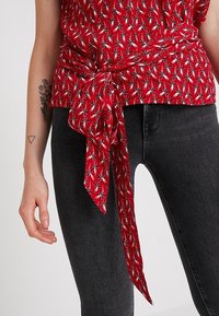 Červený tričko s krátkým rukávem a uzlem v pase, se vzorem hvězdného výbuchu v bílé barvě, ve spojení s černými upnutými džíny.