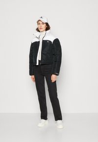 Giacca trapuntata bianca e nera, tagliata, con colletto alto e texture a piumino. Abbinata a jeans neri e sneakers bianche, indossata con un cappellino coordinato.