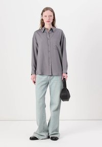 Camisa cinza de botão com um bolso, combinada com jeans flare azul claro. O modelo segura uma bolsa geométrica preta. Textura de tecido suave.