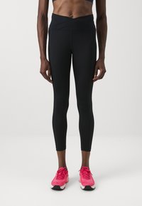 Fekete, magas derekú leggings széles, keresztezett derékrésszel, sima felülettel és testhezálló fazonnal. Élénk rózsaszín sportcipővel párosítva.