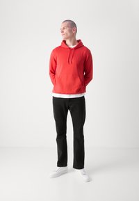 Felpa rossa con cappuccio e tasca frontale, indossata sopra una maglietta bianca, abbinata a pantaloni neri e sneakers bianche. Sfondo neutro.