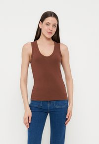 Γυναίκα με μακριά καστανά μαλλιά, που φοράει καφέ αμάνικο τοπ και μπλε τζιν, στέκεται μπροστά σε απλό φωτεινό φόντο.