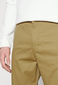 Khaki nadrág sima textúrával, egyenes szabással és kontrasztos varrással. Egy fehér, hosszú ujjú inggel párosítva, amely bemutatja a laza stílust.