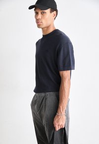 Mornarska majica s kratkimi rokavi iz mehkega materiala, kombinirana z temnimi hlačami s pinstripes vzorcem; model drži torbo, na glavi ima črn klobuk obrnjeno nazaj.