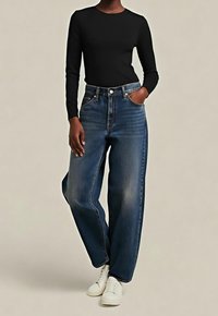 Personne portant un jean large délavé foncé, un haut noir cintré à manches longues et des baskets blanches, debout avec une main dans la poche.