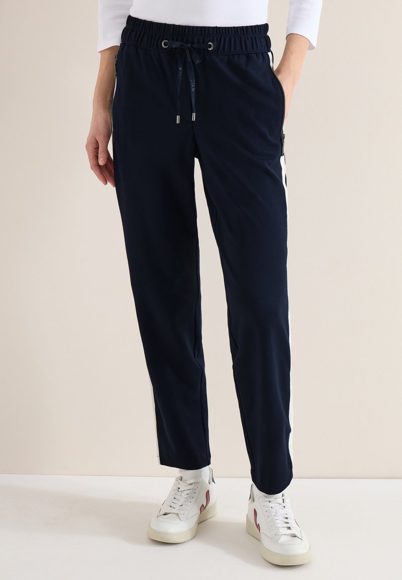 Personne portant un pantalon bleu marine à cordon avec des fermetures éclair latérales et des rayures blanches, associé à des baskets blanches et un haut blanc à manches longues.