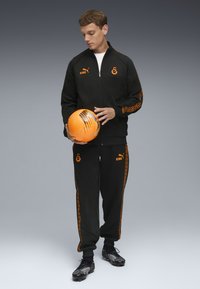 Schwarzer Trainingsanzug mit orangefarbenen Akzenten, bestehend aus einer Reißverschlussjacke und einer Hose, hält einen orangefarbenen Fußball. Schwarze Fußballschuhe mit strukturiertem Design.