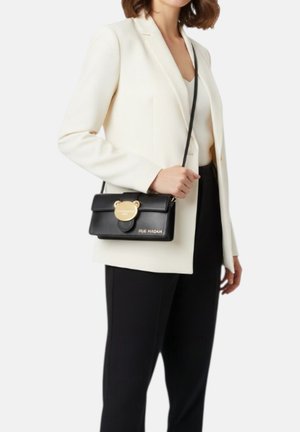 Donna con blazer bianco e pantaloni neri che tiene una borsa a tracolla nera con chiusura dorata a forma di orso e la scritta "RUE MADAM".