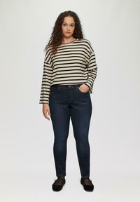 Gestreiftes, beiges und schwarzes Langarmoberteil, kombiniert mit dunkelben blauen Skinny-Jeans und schwarzen Slippern. Verfügt über eine lockere Passform und sichtbare Nähte.