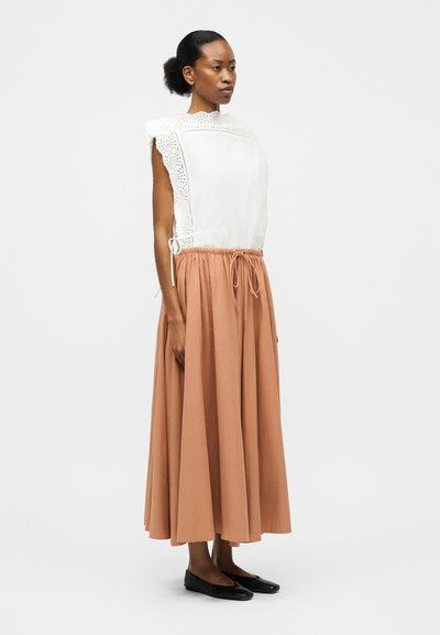 Γυναίκα με μαλλιά δεμένα πίσω, που φοράει άσπρο αμάνικο μπλουζάκι με κεντήματα και μακριά φούστα ψηλόμεση σε ροδακινί απόχρωση, καθώς και μαύρα επίπεδα παπούτσια, στέκεται πλευρικά.