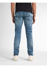 Jeans de mezclilla azul desgastado con corte recto, dos bolsillos traseros y detalles de costura sutiles, combinados con zapatillas de color claro.