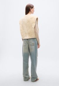 Μπεζ γιλέκο από συνθετική γούνα με φαρδιά γραμμή πάνω από ένα ανοιχτόχρωμο τοπ, συνδυασμένο με ξεθωριασμένο μπλε τζιν παντελόνι με φαρδιά πόδια και φθαρμένη λεπτομέρεια στο πίσω μέρος.