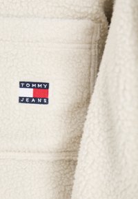 Chaqueta de forro polar color crema con un pequeño logo bordado de Tommy Jeans en rojo, blanco y azul en el bolsillo del pecho. Textura suave.