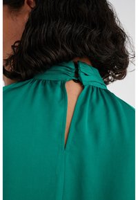 Top in tessuto verde con dettaglio arricciato sul collo, caratterizzato da una piccola apertura a chiave sul retro fissata da un bottone. Texture liscia.