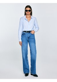 Sieviete melnās saulesbrillēs, gaiši zilā pogātā kreklā, augsta vidukļa zilās džinsās, melnā jostas siksnā un melnās smailās kurpēs, stāv ar vienu roku kabatā.