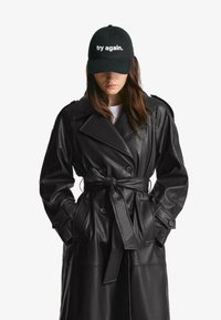 Svart konstläder trenchcoat med belagd midja, dubbelknäppt design, axelklaffar och sidofickor, kombinerad med en svart keps.