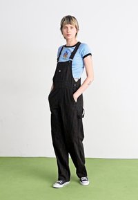 Schwarz gestreifte Latzhose aus Stoff mit Seitentaschen, getragen über einem blauen T-Shirt mit Grafikdesign und ergänzt durch schwarze Sneakers.