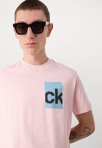 Rozā kokvilnas t-krekls ar zilu taisnstūra grafiku, kas satur treknu melnu tekstu "ck." Modelis valkā melnas kvadrātveida brilles, uzsverot minimālisma stilu.