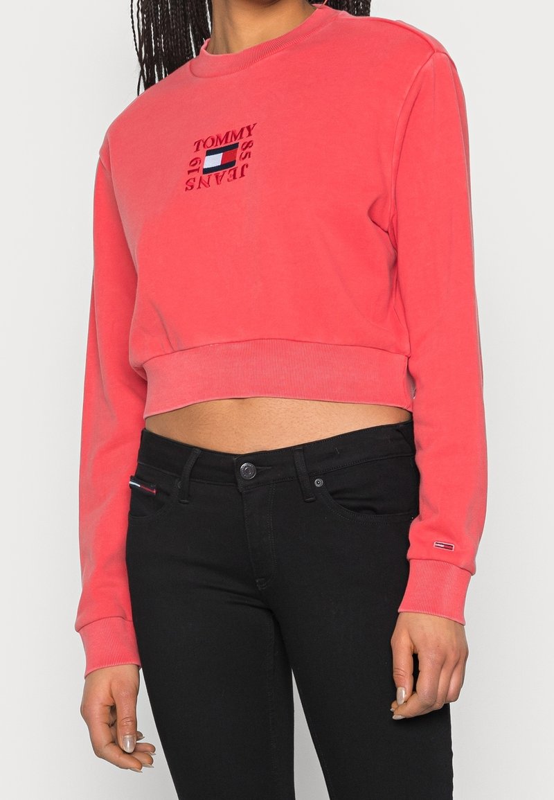 Femme portant un sweat-shirt court corail avec le logo "Tommy Jeans" et un jean taille haute noir, debout devant un fond uni.