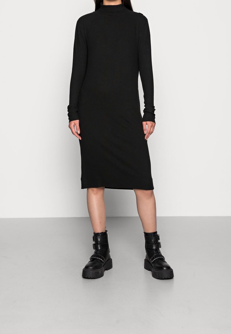 Robe noire à manches longues côtelée avec un col montant, longueur au genou, associée à des bottes noires épaisses avec des brides à boucles. Texture lisse, coupe ajustée.