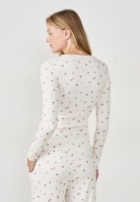 Maglietta a maniche lunghe color crema con un motivo floreale che presenta funghi e foglie. Realizzata in morbido tessuto a coste con un design aderente e scollatura tonda.