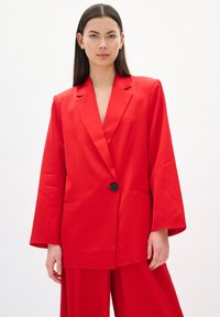 Blazer rojo con mangas largas, solapas notched, cierre de un único botón negro y bolsillos laterales, hecho de una tela suave.
