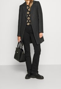 Cappotto di lana grigio, top a pois neri, pantaloni neri aderenti, borsa nera e scarpe slip-on nere. Texture liscia, vestibilità sartoriale e hardware minimale.