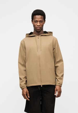 Mies, joka pukeutunut beigeen hupulliseen vetoketjutakkiin ja mustiin shortseihin, seisoo yksinkertaisen valkoisen taustan edessä.