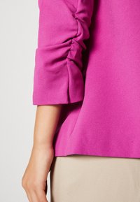 Fuchsia tröja med rynkade ärmar, mjuk textur, rundad fåll och en skräddarsydd passform, tillsammans med en ljusbeige kjol under.