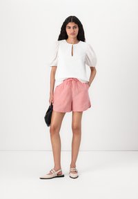 Blouse blanche texturée avec des manches bouffantes et un détail en forme de trou de clé, associée à un short rayé rouge et blanc et des chaussures plates beiges. Sac noir à la main.