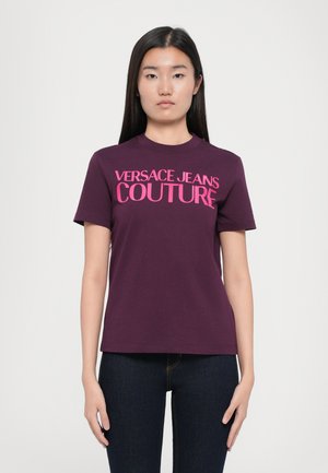 Lilla bomulls t-skjorte med korte ermer som har stor rosa "VERSACE JEANS COUTURE" tekst på forsiden. Avslappet passform, rund halsutringning.