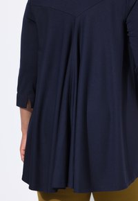 Löyhä tummansininen tunika, jossa on poimutettu selkä, kolme neljännestä hihaa ja pienet sivureiät hihansuiden lähellä. Liukas, kevyt kangas.