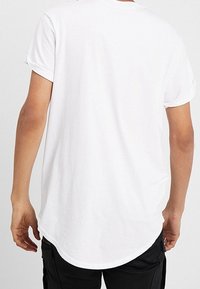 Person som bär en enkel vit t-shirt med kort ärm, sedd bakifrån, tillsammans med svarta byxor.